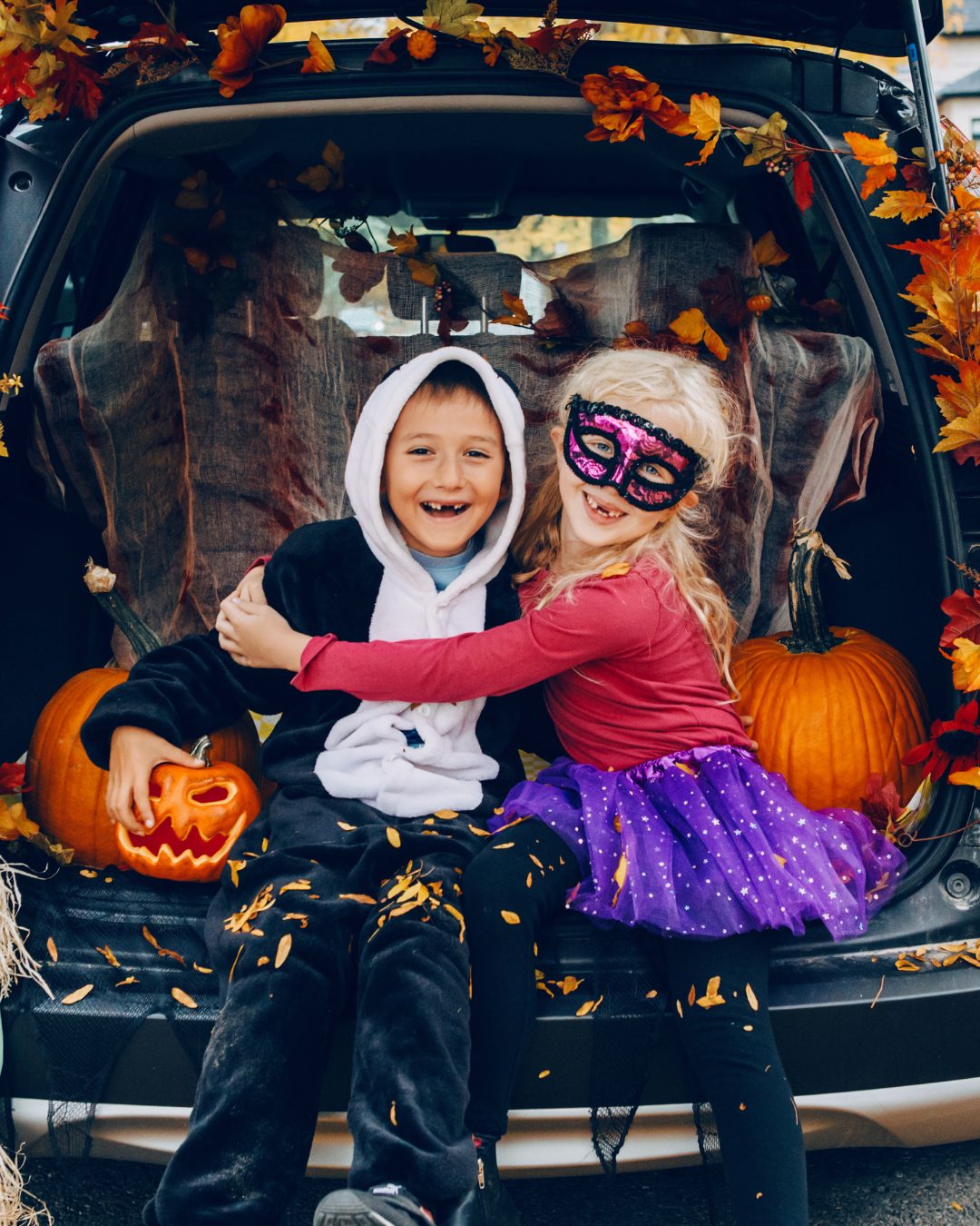 Two kids in costume laughing in the open trunk of a car decorated with fall leaves