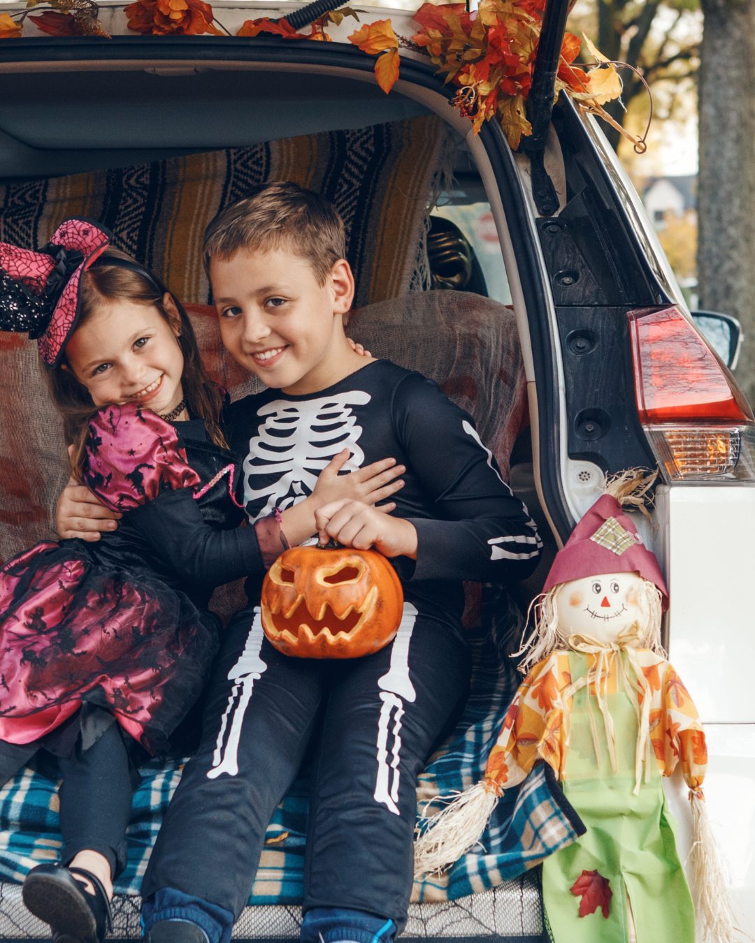 A brother and sister in costumes sitting in a decorated trunk next to a scarecrow